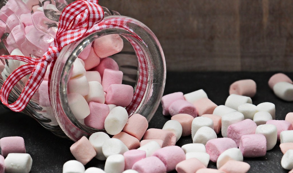 Open jar of pink and white marshmallows on its side with marshmallows falling out onto the table
